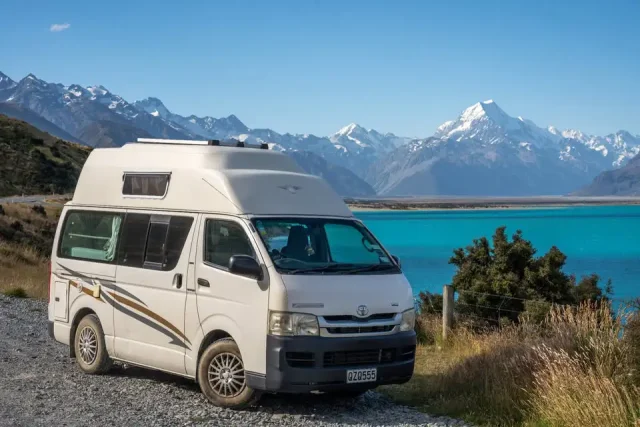 A white Toyota campervan parked beside a turquoise lake with snow-capped mountains in the background, New Zealand.